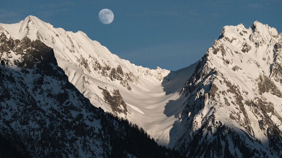 Schneebedeckte Berggipfel mit aufgehendem Mond am klaren Himmel. / Snow-covered mountain peaks with rising moon in a clear sky.