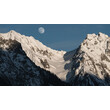 Schneebedeckte Berggipfel mit aufgehendem Mond am klaren Himmel. / Snow-covered mountain peaks with rising moon in a clear sky.