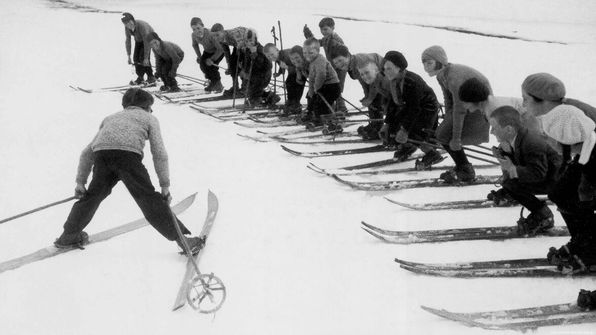 Skilehrer demonstriert Technik vor einer Reihe von Schülern im Schnee. / Ski instructor demonstrates technique to a row of learners on the slope.