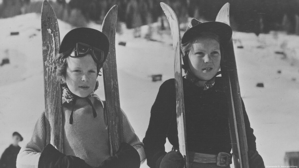 Zwei Mädchen mit Skiern im Schnee, traditionell gekleidet. / Two girls with skis in the snow, dressed traditionally.