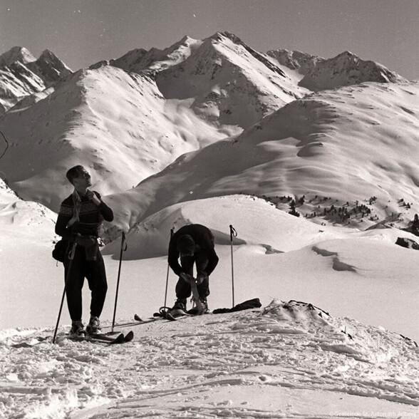 Two ski tourers in vintage gear on a snowy ridge near St. Anton am Arlberg, framed by majestic winter peaks in the background.