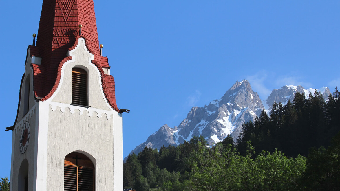 Weißer Kirchturm mit rotem Dach vor hoch aufragendem Bergmassiv. / White church tower with red roof set against towering mountain range.