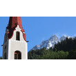 Weißer Kirchturm mit rotem Dach vor hoch aufragendem Bergmassiv. / White church tower with red roof set against towering mountain range.