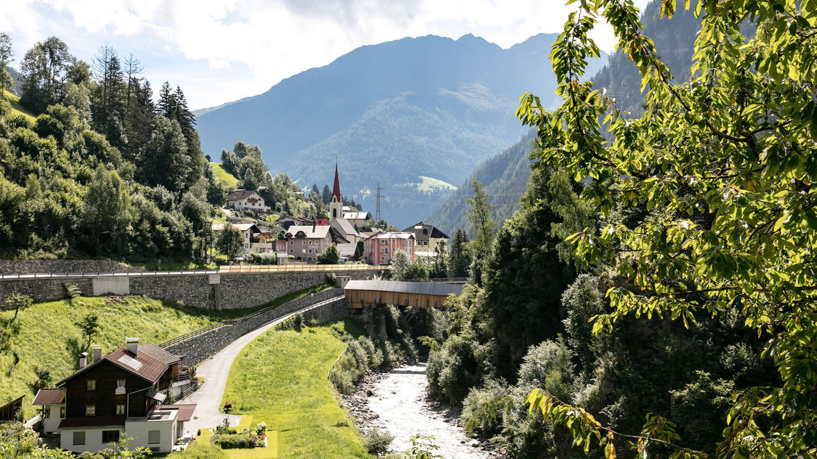 Strengen im Sommer mit grünen Wiesen, Fluss und Kirche vor alpiner Kulisse. / Summer in Strengen with green meadows, river, and church against alpine backdrop.