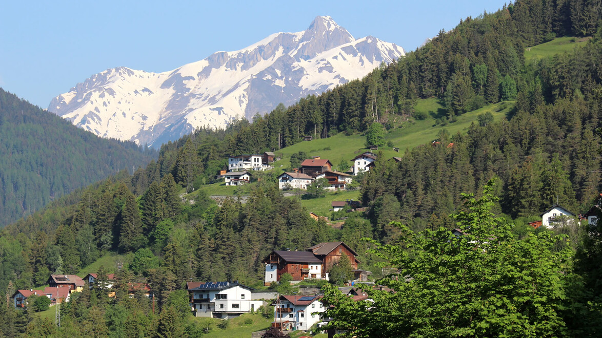 Bergdorf mit verstreuten Häusern an grüner Hanglage und schneebedecktem Gipfel im Hintergrund. / Mountain village with scattered hillside houses and snowy summit in background.