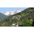Bergdorf mit verstreuten Häusern an grüner Hanglage und schneebedecktem Gipfel im Hintergrund. / Mountain village with scattered hillside houses and snowy summit in background.