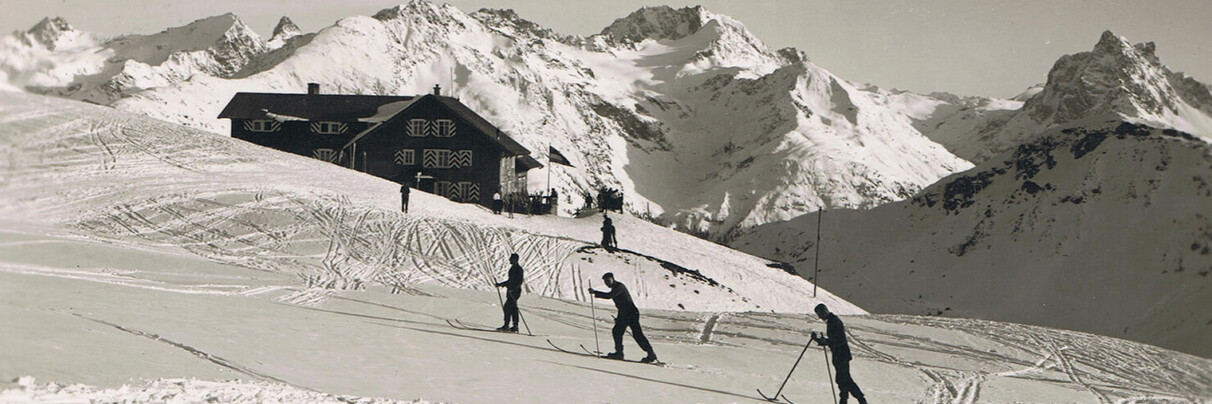 “Historic winter scene with ski tourers near a mountain hut in the Alps above St. Anton am Arlberg, surrounded by snow-covered peaks and open slopes.