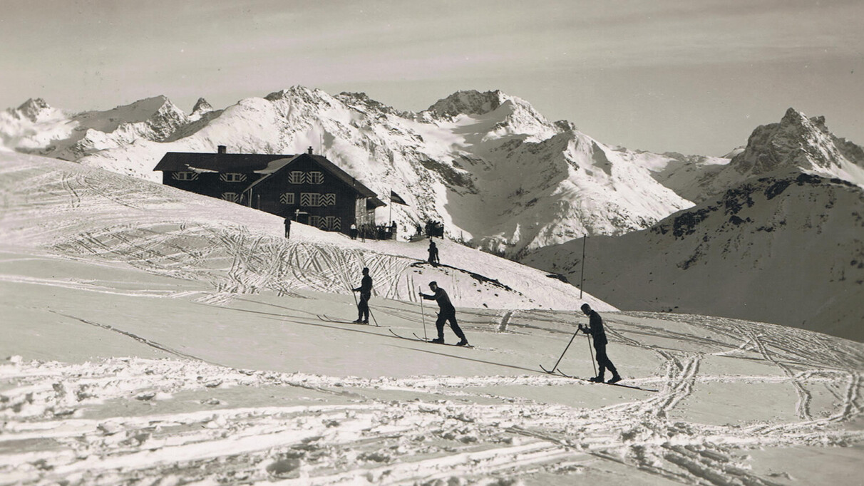 “Historic winter scene with ski tourers near a mountain hut in the Alps above St. Anton am Arlberg, surrounded by snow-covered peaks and open slopes.