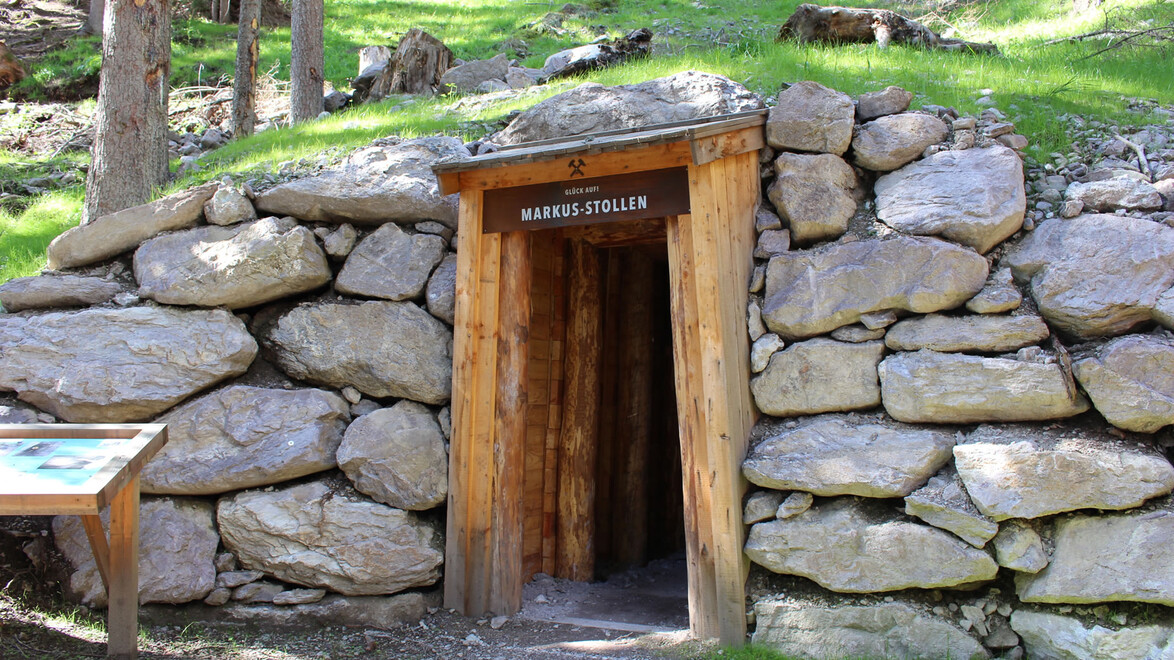 Kleiner Stolleneingang im Wald mit Holzverkleidung und Schlägel-und-Eisen-Symbol. / Small wooden mine entrance in the forest with hammer and pick symbol.