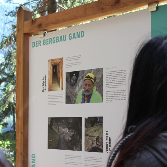 Outdoor information board about the historic Gand mining area in St. Anton am Arlberg, featuring photos of a tunnel, a geologist with a helmet, and hikers on the themed trail.