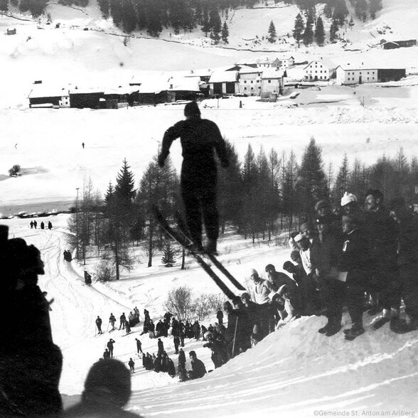 Historische Skisprungveranstaltung in St. Anton am Arlberg: Ein Skispringer in der Luft vor winterlicher Kulisse, umringt von zahlreichen Zuschauer:innen auf der verschneiten Schanze.