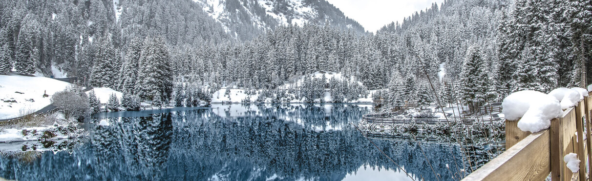 Snow-covered winter landscape with a deep blue Verwall Lake surrounded by dense fir forests near St. Anton am Arlberg – wooden railing with snow in the foreground, trees mirrored in the calm water.