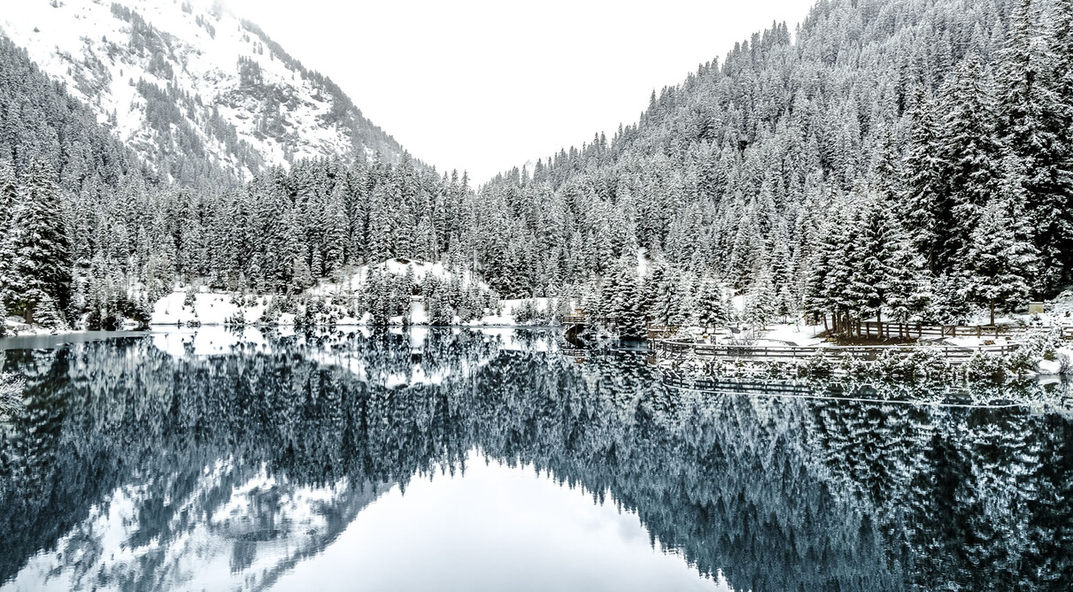 Snow-covered winter landscape with a deep blue Verwall Lake surrounded by dense fir forests near St. Anton am Arlberg – wooden railing with snow in the foreground, trees mirrored in the calm water.