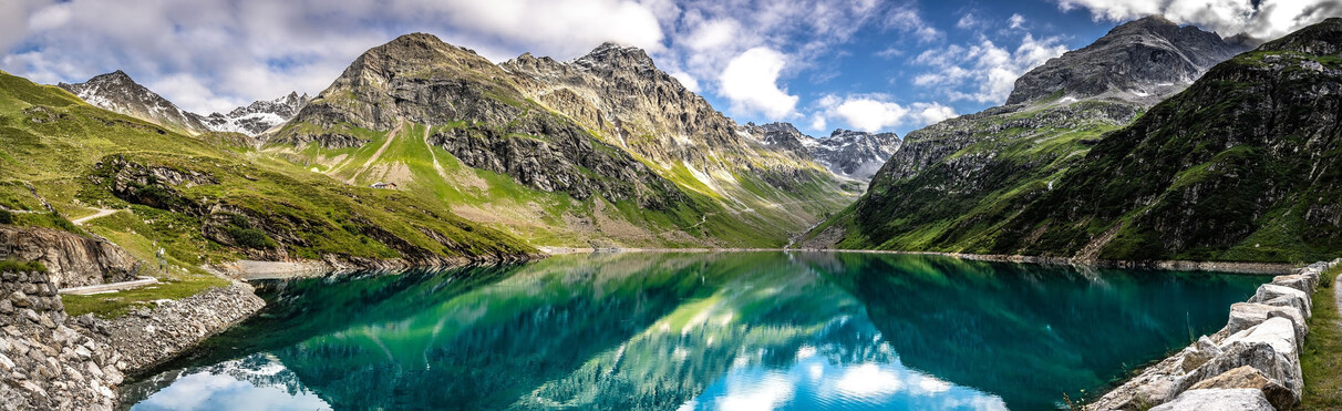 Crystal-clear Kartell reservoir nestled in the Tyrolean Alps near St. Anton am Arlberg – green meadows, rocky peaks and cloud reflections in turquoise water.