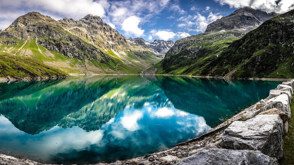 Crystal-clear Kartell reservoir nestled in the Tyrolean Alps near St. Anton am Arlberg – green meadows, rocky peaks and cloud reflections in turquoise water.