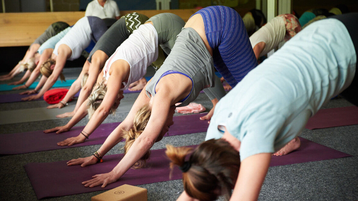 Mehrere Personen üben im Yogaraum gleichzeitig die Haltung „herabschauender Hund“. — Several people perform the downward-facing dog pose in a yoga room.