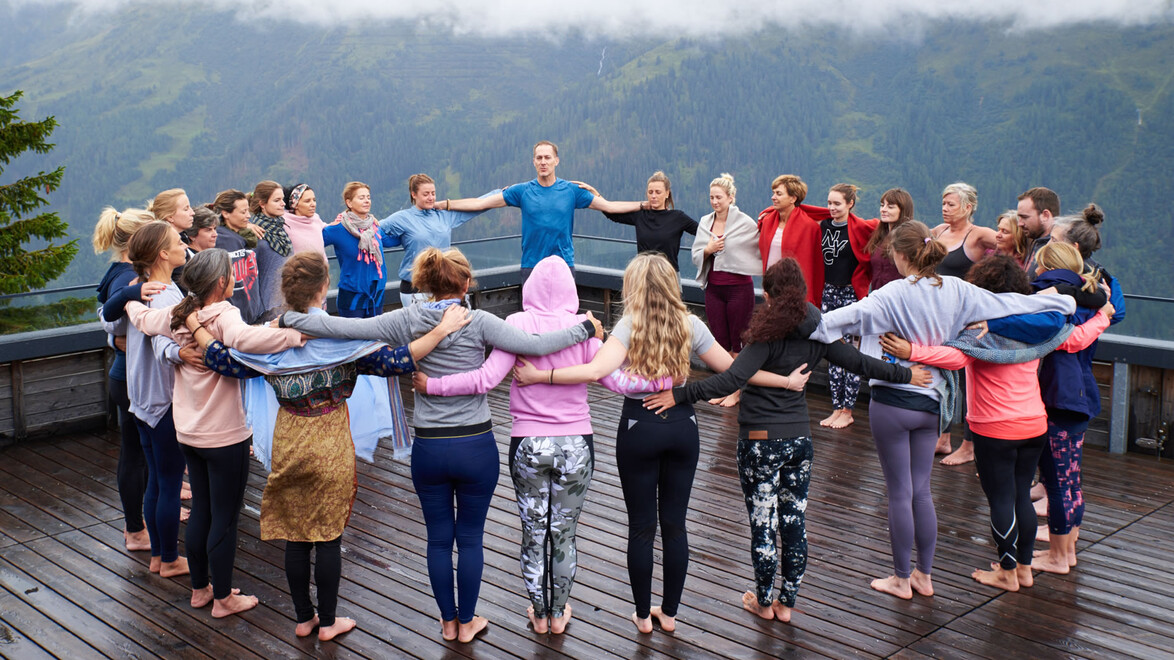 Teilnehmende stehen im Kreis auf einer Terrasse und halten sich an den Schultern. — Participants stand in a circle on a terrace, holding each other's shoulders.