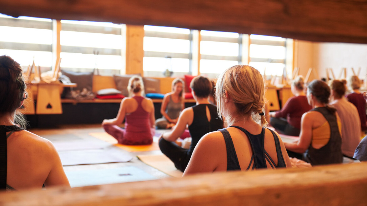 Mehrere Personen sitzen im Yogaraum auf Matten im Schneidersitz. — A group of people sits cross-legged on yoga mats in an indoor yoga room.