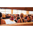 Mehrere Personen sitzen im Yogaraum auf Matten im Schneidersitz. — A group of people sits cross-legged on yoga mats in an indoor yoga room.
