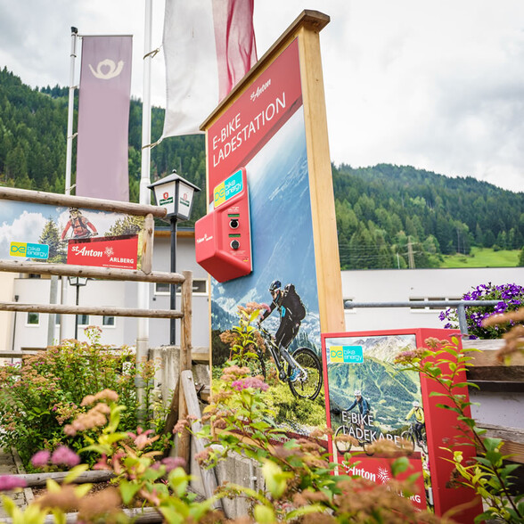 An e-bike charging station with a red housing stands next to signs and flowers. In the background are woods and buildings.
