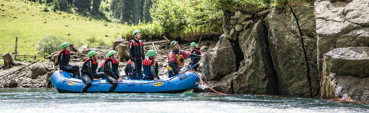 Group of kids in a blue raft learning safely near a rocky riverbank under sunny skies.