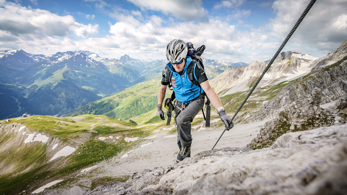 Ein Bergsteiger klettert mit Helm und Klettersteigset an einem Drahtseil entlang auf einem felsigen Pfad mit Panoramaausblick. / A climber with helmet and via ferrata gear ascends a rocky trail secured by a steel rope, with panoramic mountain views.