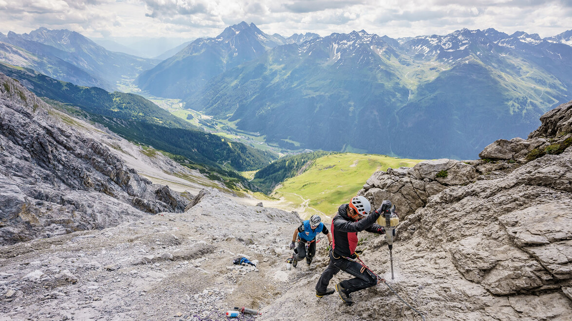 Zwei Kletterer steigen über steiniges Gelände in einem gesicherten Klettersteig mit Weitblick auf grüne Täler. / Two climbers ascend rocky terrain on a secured via ferrata with a wide view of green valleys.