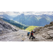 Zwei Kletterer steigen über steiniges Gelände in einem gesicherten Klettersteig mit Weitblick auf grüne Täler. / Two climbers ascend rocky terrain on a secured via ferrata with a wide view of green valleys.