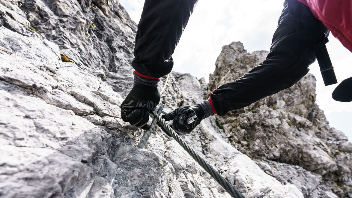 Nahaufnahme eines Klettersteig-Abschnitts mit Fokus auf Schuhwerk und Drahtseil zur Sicherung. / Close-up of a via ferrata section showing climbing shoes and steel rope for safety.