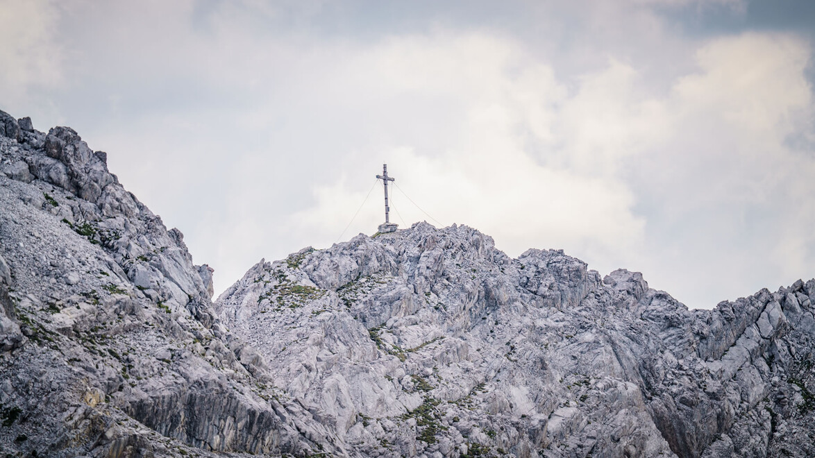 Ein Gipfelkreuz steht auf einem felsigen Berggrat unter bewölktem Himmel. / A summit cross stands on a rocky mountain ridge under a cloudy sky.