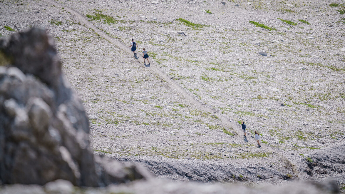 Wanderer bewegen sich über einen felsigen Hang mit Drahtseilsicherung, aus der Ferne fotografiert. / Hikers move across a rocky slope secured with steel ropes, captured from a distance.