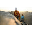 Zwei Kletterer auf einem schmalen Felsgrat bei Sonnenaufgang mit Seilsicherung. / Two climbers on a narrow rock ridge at sunrise, secured with rope.