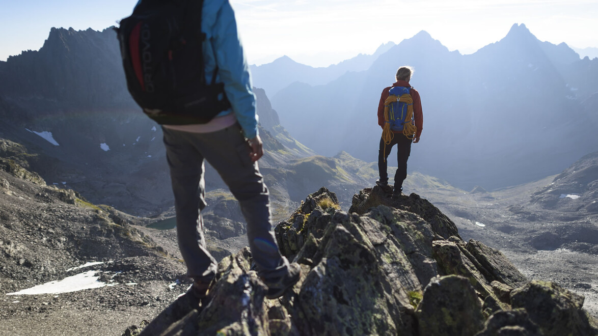 Zwei Bergsteiger stehen auf einem felsigen Gipfel mit Aussicht über ein zerklüftetes Bergmassiv. / Two mountaineers stand on a rocky summit with views over a rugged mountain range.