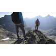 Zwei Bergsteiger stehen auf einem felsigen Gipfel mit Aussicht über ein zerklüftetes Bergmassiv. / Two mountaineers stand on a rocky summit with views over a rugged mountain range.