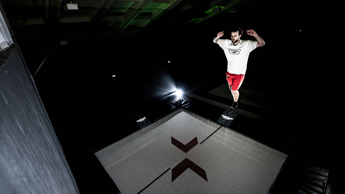 Ein Sportler springt mit ausgebreiteten Armen über einen großen Airbag im Dunkeln. / An athlete jumps with outstretched arms over a large airbag in the dark.