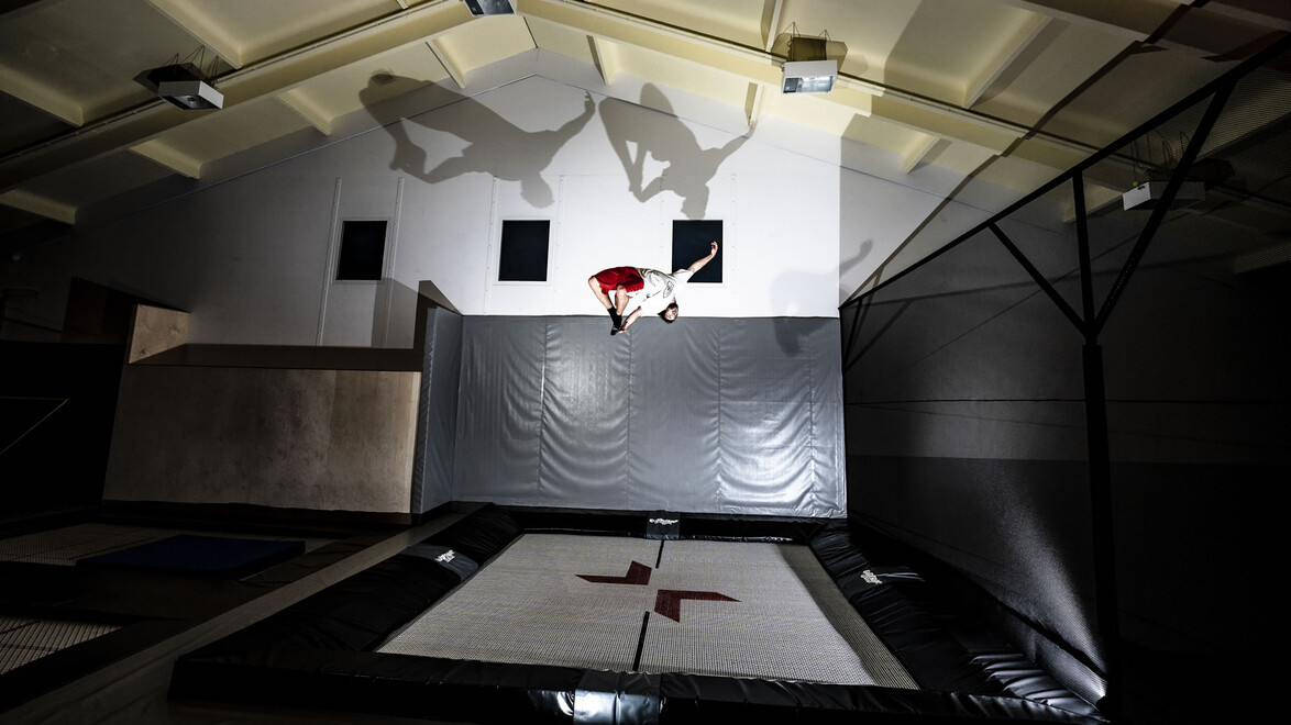 Zwei Personen springen vor einem Basketballkorb – ihre Schatten werden an die Wand projiziert. / Two people jump in front of a basketball hoop – their shadows are cast on the wall.
