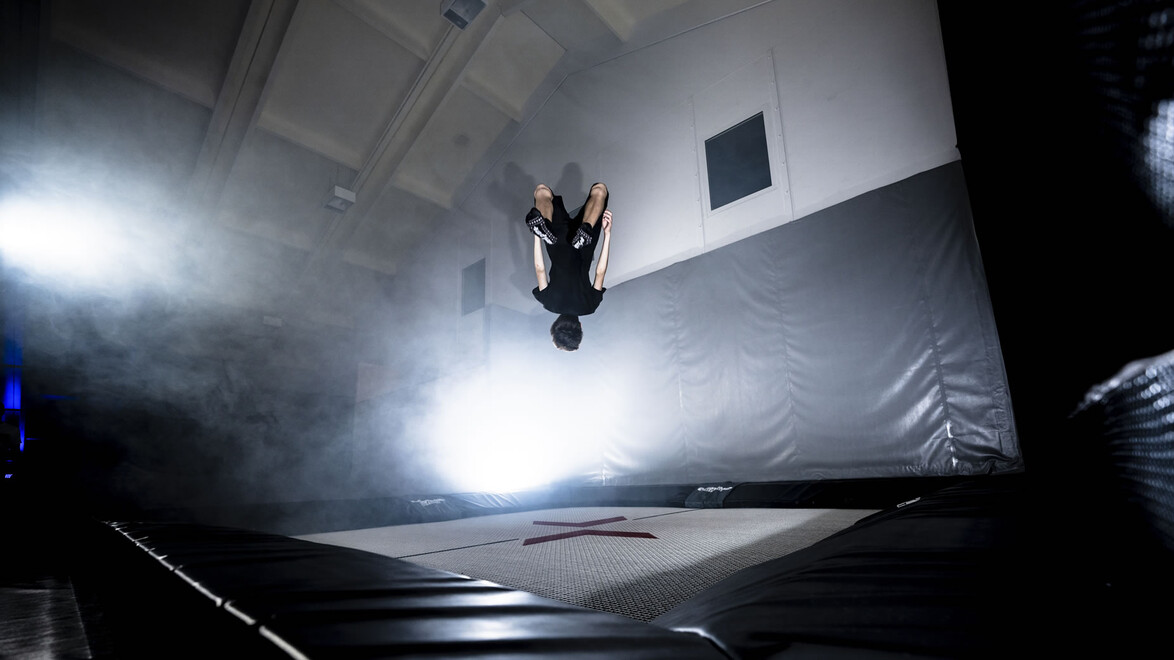Ein Freestyler macht einen Rückwärtssalto, umgeben von Licht und Nebel auf einer Indoor-Fläche. / A freestyler performs a backflip surrounded by light and fog on an indoor surface.