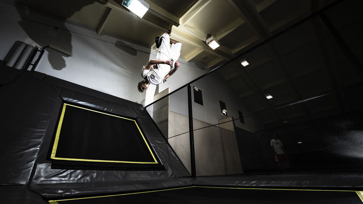 Ein junger Sportler springt mit Schwung von einem schwarzen Trampolin in einer dunklen Halle. / A young athlete jumps dynamically from a black trampoline in a dark indoor hall.