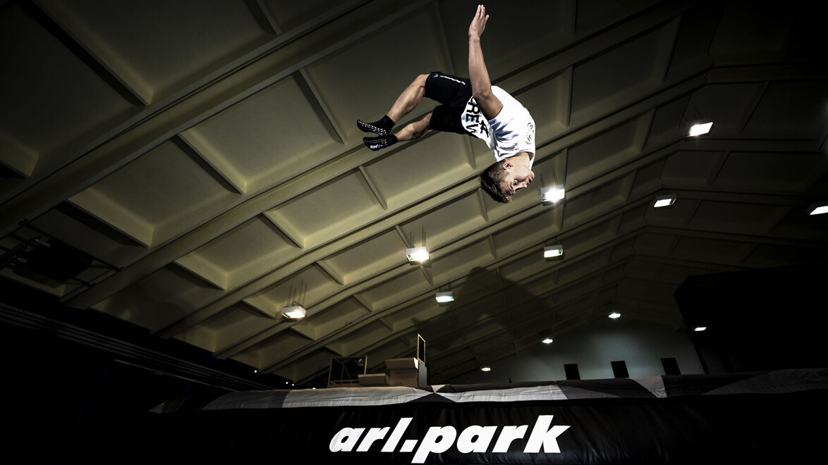 Ein Jugendlicher zeigt einen Rückwärtssalto in der beleuchteten Trampolinhalle arl.park. / A teenager performs a backflip in the illuminated trampoline hall arl.park.