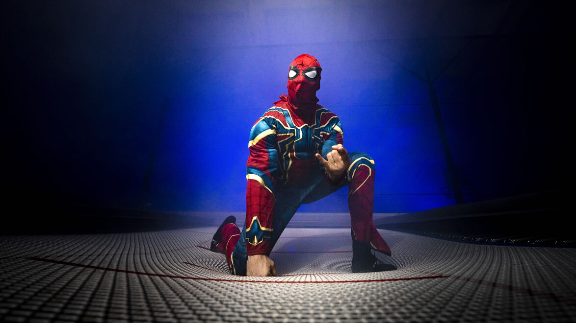 Ein als Superheld verkleideter Akrobat posiert auf einem beleuchteten Sprungfeld. / An acrobat dressed as a superhero poses on a lit jump platform.