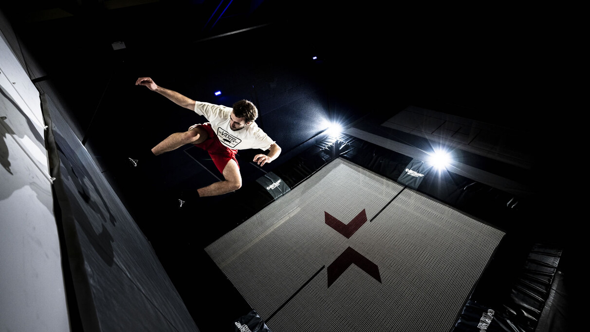 Mann springt auf einem Trampolin im Dunkeln mit professioneller Beleuchtung. Es trägt eine kurze rote Sporthose und ein weißes T-Shirt, er befindet sich gerade in eine Hocke in der Luft schwebend.