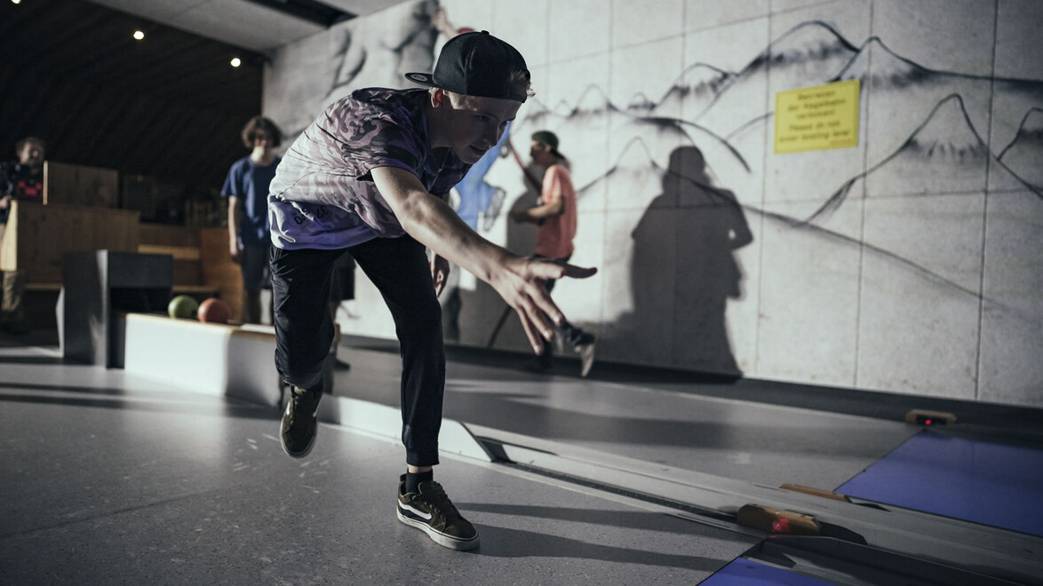 Jugendlicher beim Wurf, bunt beleuchtete Kegelbahn mit Wandmalerei / Teenager bowling, colorfully lit lane with mural on the wall