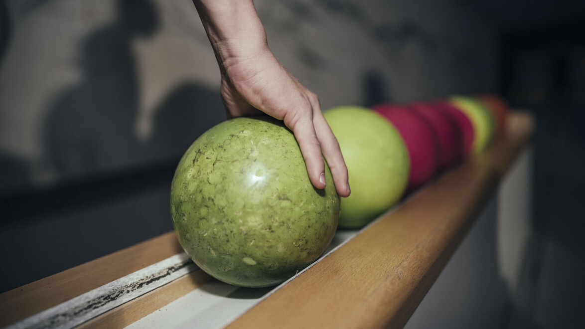 Nahaufnahme einer Hand, die eine grüne Kugel aufnimmt, mehrere Kugeln in Reihe / Close-up of a hand grabbing a green bowling ball, several balls in a row
