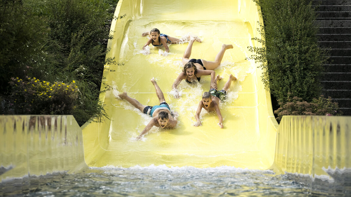 Vier Kinder rutschen lachend nebeneinander auf einer gelben Wasserbahn ins Becken / Four children slide down a yellow waterslide together, laughing and splashing into the pool