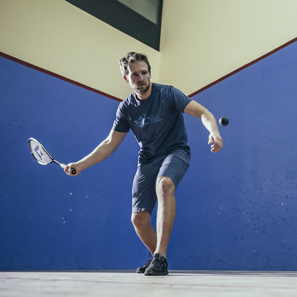 A man plays squash in an indoor court with blue walls, focused on the ball while holding his racket.