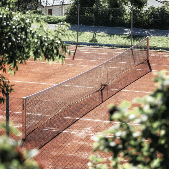 Red clay tennis court with net in summer sunlight, framed by green trees and hedges.