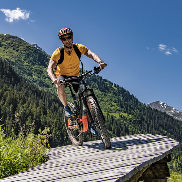 A man in a yellow T-shirt rides his mountain bike on the Northshore Trail over a wooden footbridge in the middle of the green mountain landscape of Verwalltal in St. Anton am Arlberg.