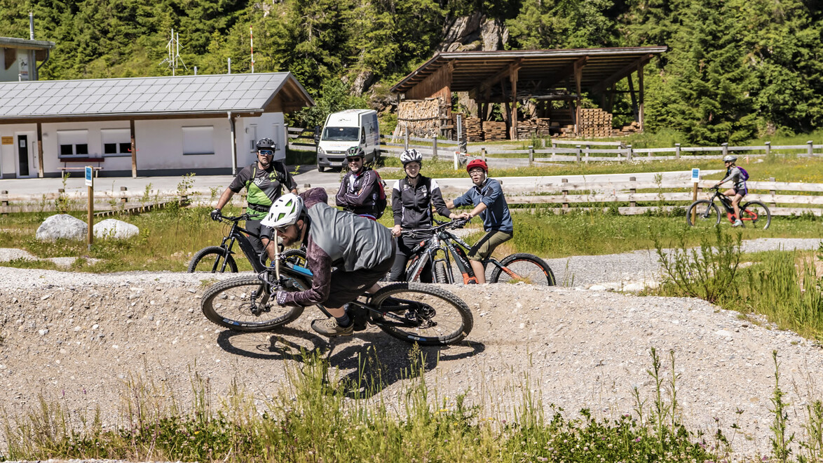 Gruppe von Radfahrer:innen wartet beim Pumptrack, eine Person fährt aktiv / Group of cyclists waiting at the pump track, one person riding actively