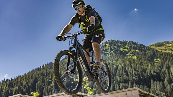 A man rides an e-mountain bike over a wooden ramp, with green forests and blue sky in the background.