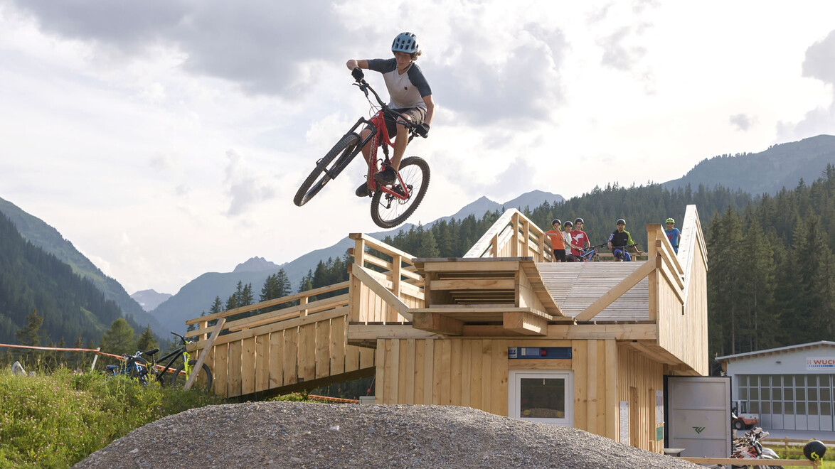 Biker springt über Erdhügel vor Holzgebäude im Bikepark / Biker jumps over dirt hill in front of wooden building at bike park